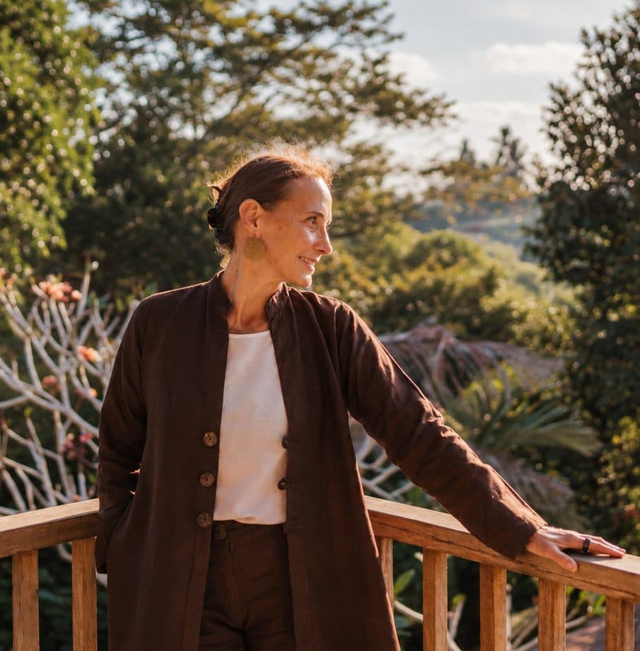 Christiane in braunem Blazer auf einem Balkon im Grünen, Blick in die Ferne.
