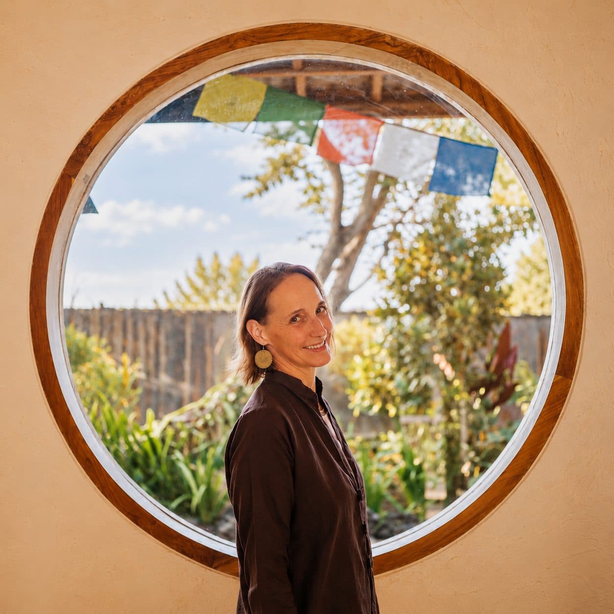 Christiane steht lächelnd vor einem runden Fenster, dahinter Garten und Gebetsfahnen.