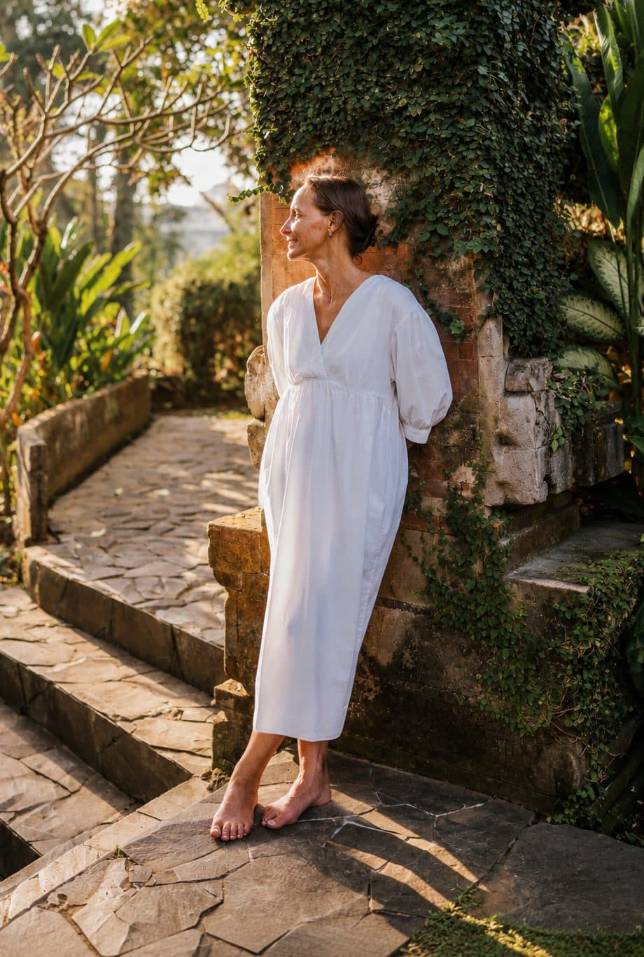 Christiane lehnt in weissem Kleid an einer efeubewachsenen Steinmauer, Blick zur Seite.