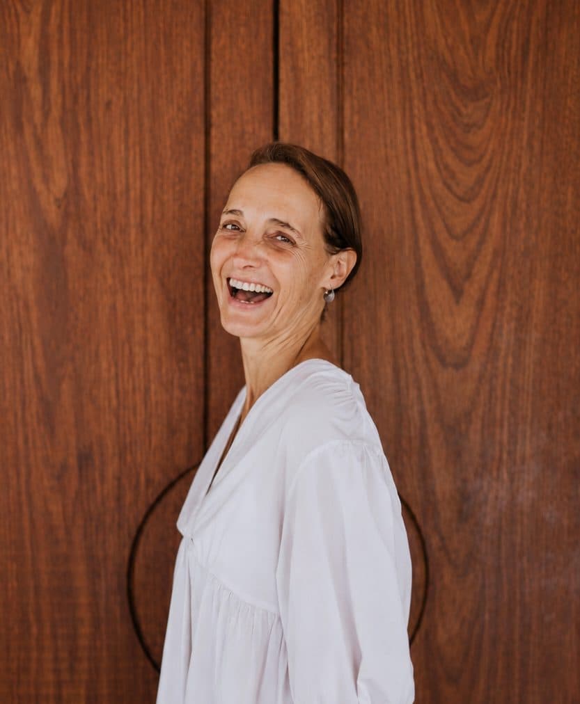 Christiane lacht vor einer Holzwand, die Hände in den Taschen.